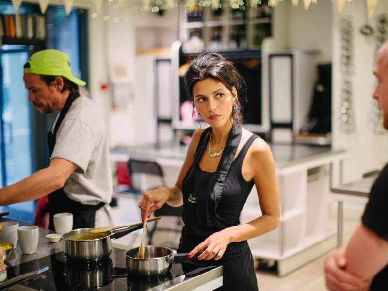 Two people cooking whilst listening to a teacher at a cooking class